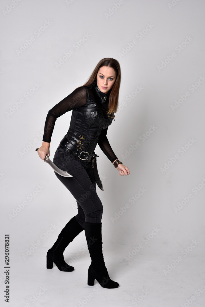 full length portrait of a pretty brunette woman wearing black leather fantasy costume. Standing pose, holding a dagger on a studio background. full length portrait of a pretty brunette woman wearing black leather fantasy costume. Standing pose, holding a dagger on a studio background.