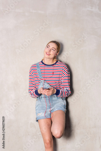 happy blond girl listening to music with earphones and smartphone by wall