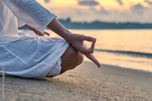Woman in white clothes and a turban sits on a sandy beach at dawn and practices yoga