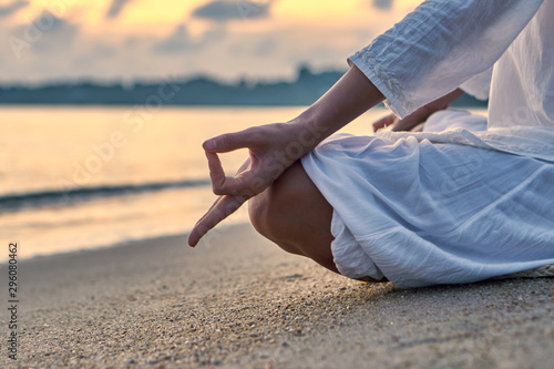 Woman in white clothes and a turban sits on a sandy beach at dawn and practices yoga