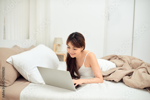 Smiling Asian young woman using laptop in bed at home
