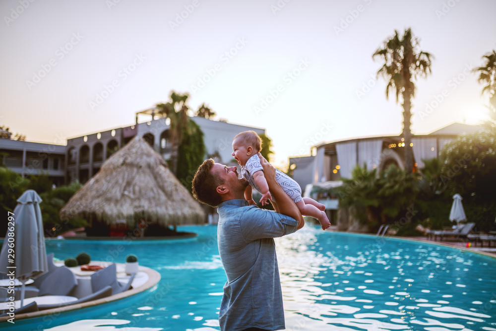 Side view of happy Caucasian dad lifting his adorable 6 months old son while standing next to swimming pool at sunset.