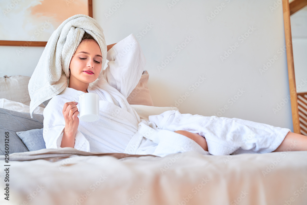 Image of happy woman in white bathrobe with mug of tea in her hands lying on bed. Image of happy woman in white bathrobe with mug of tea in her hands lying on bed.