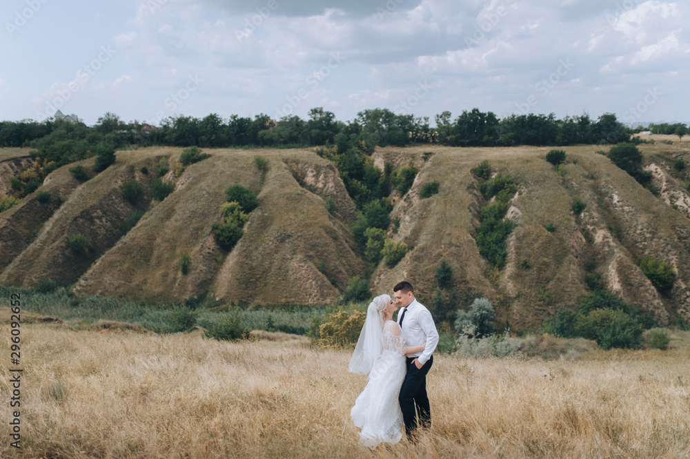 Stylish groom in a white shirt hugs a beautiful and smiling blonde bride against the background of nature, hills. Wedding portrait of lovers newlyweds. Photography and concept. Stylish groom in a white shirt hugs a beautiful and smiling blonde bride against the background of nature, hills. Wedding portrait of lovers newlyweds. Photography and concept.