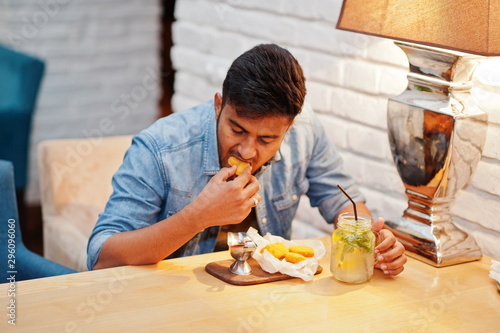 Portrait of handsome successful bearded south asian  young indian freelancer in blue jeans shirt sitting in cafe with chicken nuggets and lemonade.