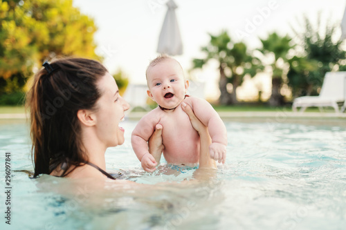 Cheerful Caucasian 6 months old baby boy learning how to swim at swimming pool. Mother holding her son. First time at pool concept.