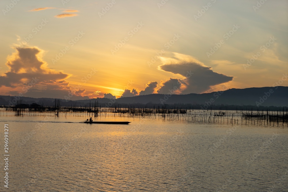 Sunrise view over the lake with small fisherman hut in the middle, fisherman sailing small boat to check fishing trap with morning light and mountains in background.