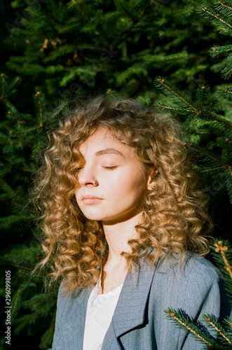 A young adult beautiful girl with blond curly hair stands on a green background with her eyes closed. The face of a young girl bathed in warm sunlight