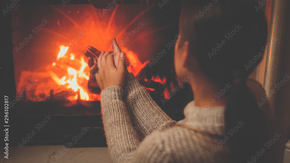 Toned photo of young woman in woolen sweater sitting by the burning fireplace and warming hands Toned photo of young woman in woolen sweater sitting by the burning fireplace and warming hands