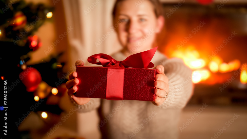 Closeup image of young woman showing red Christmas gift box in camera while sitting next to Xmas tree and burning fireplace Closeup image of young woman showing red Christmas gift box in camera while sitting next to Xmas tree and burning fireplace