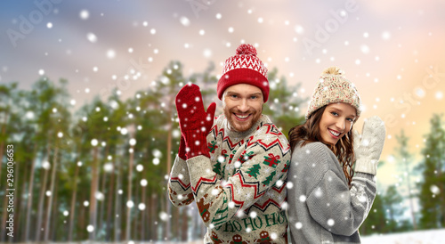 christmas, winter clothes and holidays concept - portrait of happy couple in mittens, hats and ugly sweaters over snow and forest background