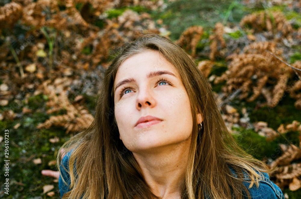 A beautiful young adult girl with blond hair and blue eyes sits on green autumn grass and moss against the background of dry yellow autumn leaves and looks up at the sky A beautiful young adult girl with blond hair and blue eyes sits on green autumn grass and moss against the background of dry yellow autumn leaves and looks up at the sky