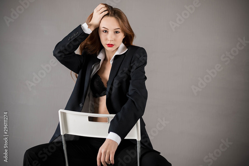 Sexy lady posing sitting on a chair, dressed in a black suit.The red-haired girl. Gray background. photo