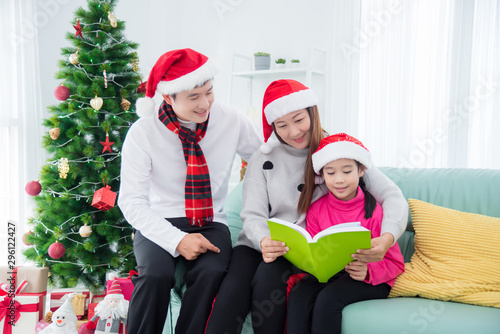 Happy asian family sitting on sofa and reading book together in room with christmas tree decoration