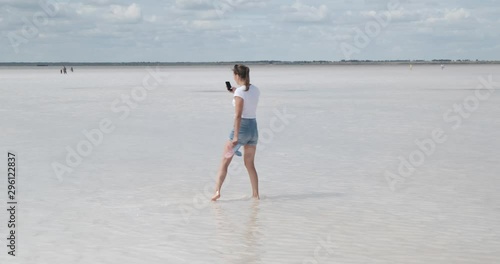 Beautiful girl in white T-shirt and shorts walks on shallows of salt treatment lake, resort abroad, Izrail, summer, huge dead lake, takes pictures of everything around on phone, walks on salt water photo