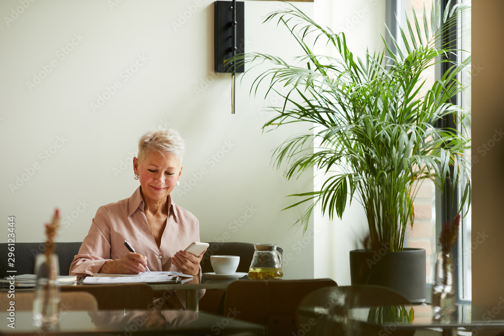 Mature smiling businesswoman using her mobile phone and making notes at the table in cafe Mature smiling businesswoman using her mobile phone and making notes at the table in cafe