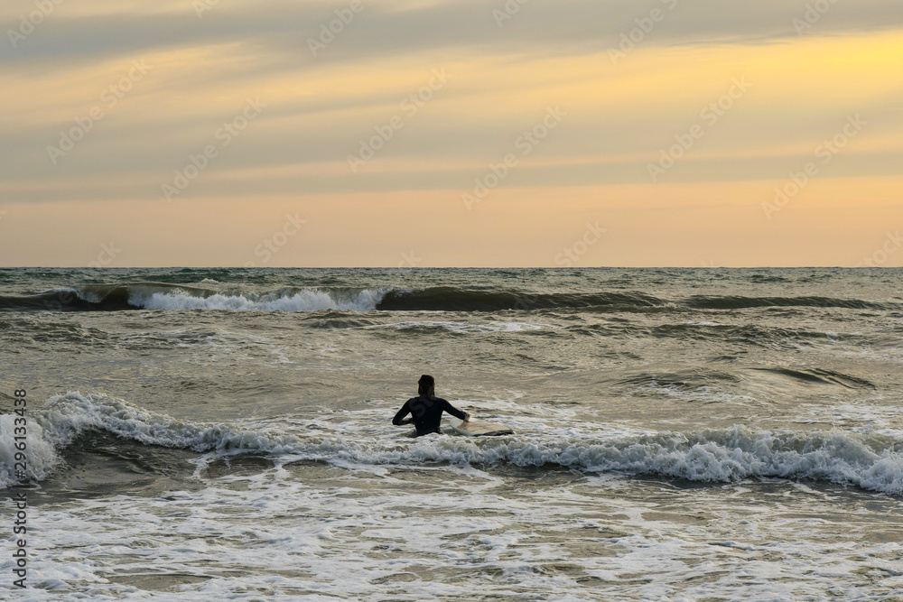Seascape with the silhouette of a surfer from behind waiting for the perfect wave on the shore at sunset, Tuscany, Italy