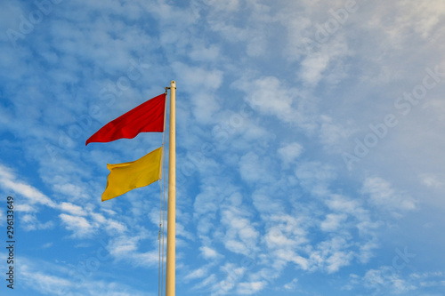 Top of a pole with a red and a yellow warning flags, indicating total absence of surveillance on the beach, against a cloudy blue sky, Italy