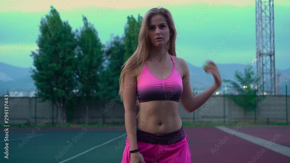 Young pretty girl plays tennis. A blonde with long hair in a pink tracksuit professionally trains tennis. The girl serves the ball. Young pretty girl plays tennis. A blonde with long hair in a pink tracksuit professionally trains tennis. The girl serves the ball.