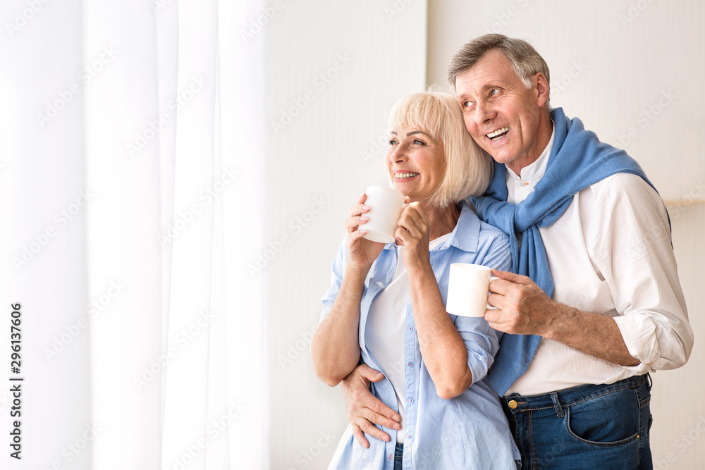 Loving senior couple standing near window and drinking tea Loving senior couple standing near window and drinking tea