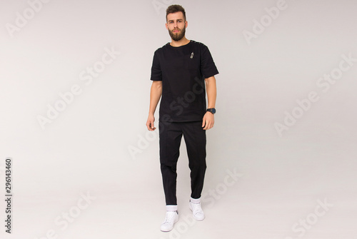 young stylish bearded nurse male in black medical costume is standing straight and smiling cute on the white wall background. medical fashion concept. free space