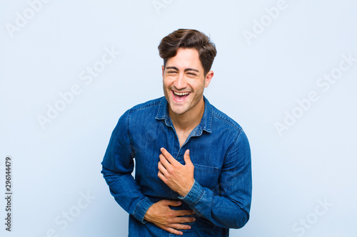 young handsome man laughing out loud at some hilarious joke, feeling happy and cheerful, having fun against blue background