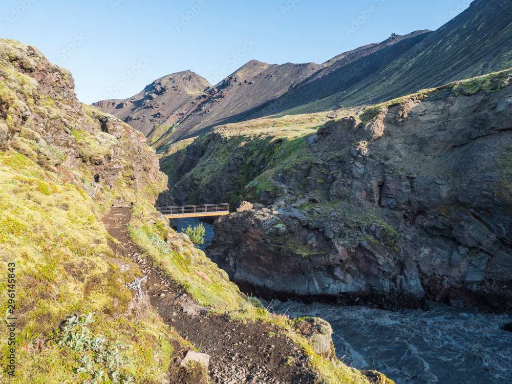 Landscape with wooden footbridge over a blue glacier river at laugavegur hiking trail in iceland. Lava formation and green hills and mountains, summer blue sky background