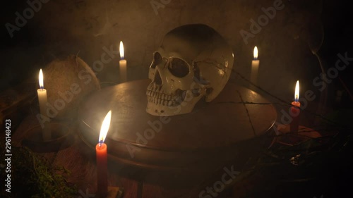 Human skull rotating on a wooden circular platform with dark gray background with candles and mystic herbs around a table with smoke
