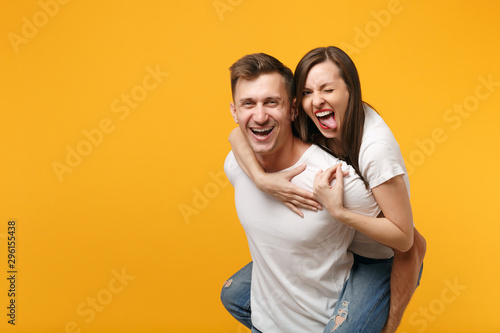 Laughing young couple two friends guy girl in white t-shirts posing isolated on yellow orange background. People lifestyle concept. Mock up copy space. Giving piggyback ride to joyful sitting on back.