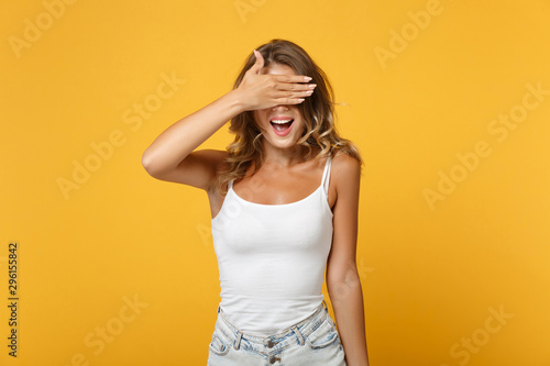 Cheerful young woman in light casual clothes posing isolated on yellow orange wall background, studio portrait. People sincere emotions lifestyle concept. Mock up copy space. Covering eyes with hand.