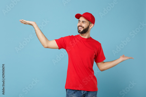 Delivery man in red uniform workwear isolated on blue wall background, studio portrait. Professional male employee in cap t-shirt print working as courier dealer. Service concept. Mock up copy space.