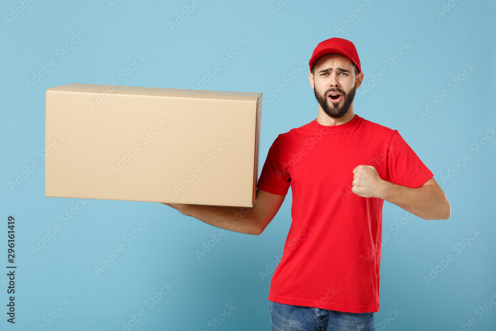 Delivery man in red uniform isolated on blue background, studio portrait. Male employee in cap t-shirt print working as courier dealer hold empty cardboard box. Service concept. Mock up copy space. Delivery man in red uniform isolated on blue background, studio portrait. Male employee in cap t-shirt print working as courier dealer hold empty cardboard box. Service concept. Mock up copy space.