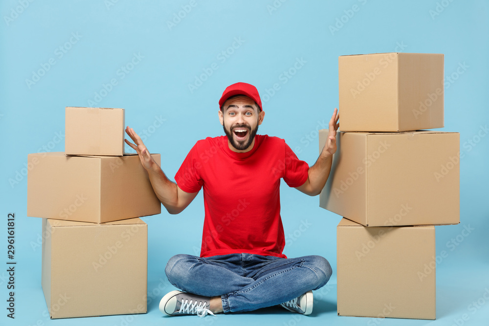 Delivery man in red uniform isolated on blue background, studio portrait. Male employee in cap t-shirt print working as courier dealer hold empty cardboard box. Service concept. Mock up copy space. Delivery man in red uniform isolated on blue background, studio portrait. Male employee in cap t-shirt print working as courier dealer hold empty cardboard box. Service concept. Mock up copy space.