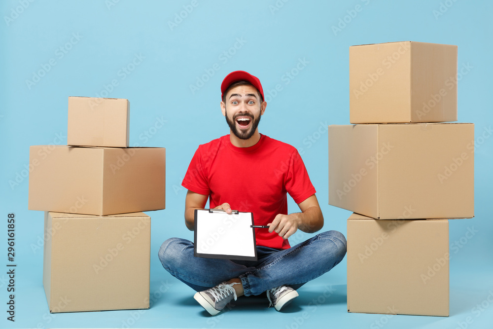 Delivery man in red uniform isolated on blue background, studio portrait. Male employee in cap t-shirt print working as courier dealer sit at empty cardboard box. Service concept. Mock up copy space. Delivery man in red uniform isolated on blue background, studio portrait. Male employee in cap t-shirt print working as courier dealer sit at empty cardboard box. Service concept. Mock up copy space.