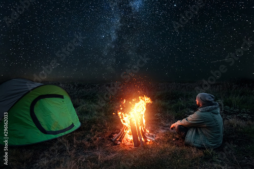 Camping man sits by the fire at night against the background of the starry sky. The concept of travel, tourism, camping.