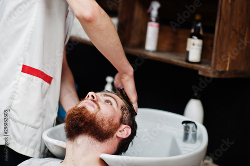 Young bearded man washing head by hairdresser while sitting in chair at barbershop. Barber soul.