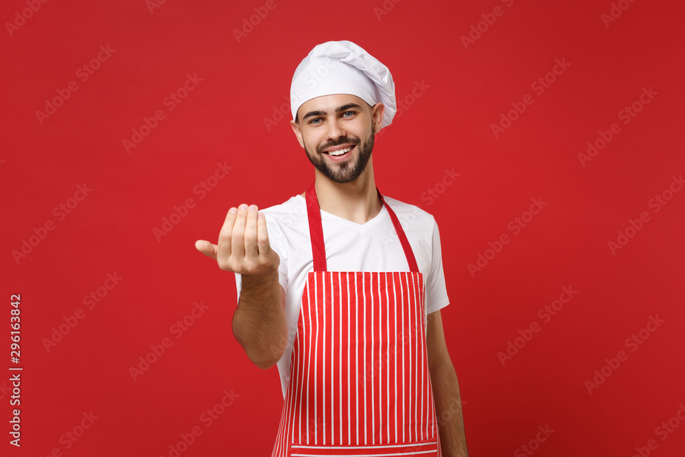 Young bearded male chef cook or baker man in striped apron white t-shirt toque chefs hat posing isolated on red background. Cooking food concept. Mock up copy space. Doing gesture like says come here. Young bearded male chef cook or baker man in striped apron white t-shirt toque chefs hat posing isolated on red background. Cooking food concept. Mock up copy space. Doing gesture like says come here.