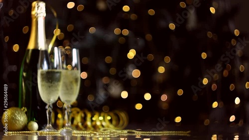 Two glasses and green bottle with champagne, golden decor, balls, serpentine are on table. Festive decorative garland, warm yellow light bulbs are blinking on background. New year, christmas mood. 