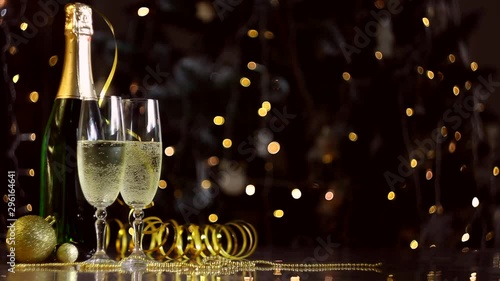 Two glasses and green bottle with champagne, golden decor, balls, serpentine are on table. Festive decorative garland, warm yellow light bulbs are blinking on background. New year, christmas mood. 