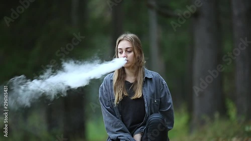 Vape teenager. Young beautiful white teenage girl in casual clothes vaping electronic cigarette on the street in the park in the autumn evening. Bad habit. Vaping activity.