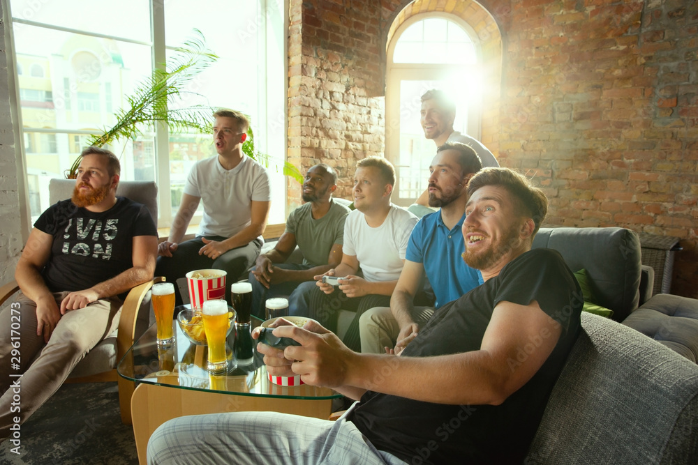 Group of excited friends playing video games at home. Caucasian male gamers or fans spending time and having fun together at home. Emotional watching gameplay. Modern technologies, friendship, weekend
