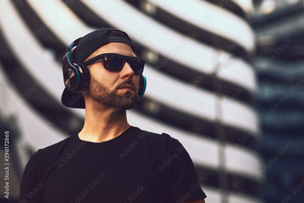 Portrait of a young bearded man with a black hat and sunglasses in black t-shirt in the city Portrait of a young bearded man with a black hat and sunglasses in black t-shirt in the city