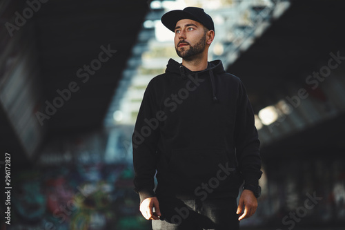 Portrait of a young bearded man with a black hat ain black hoodie in the city