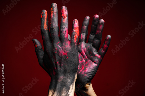 Men's hands in black, pink and red paint showing a clapping gesture on a dark background. Right hand in front of left. Close-up studio photo