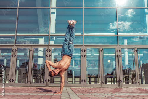 dancer in jump stands on one arm, acrobat actor break dance, hip-hop. In summer city, background glass windows clouds lifestyle, young dancer, fitness workout breakdancer jeans sneakers. Free space.