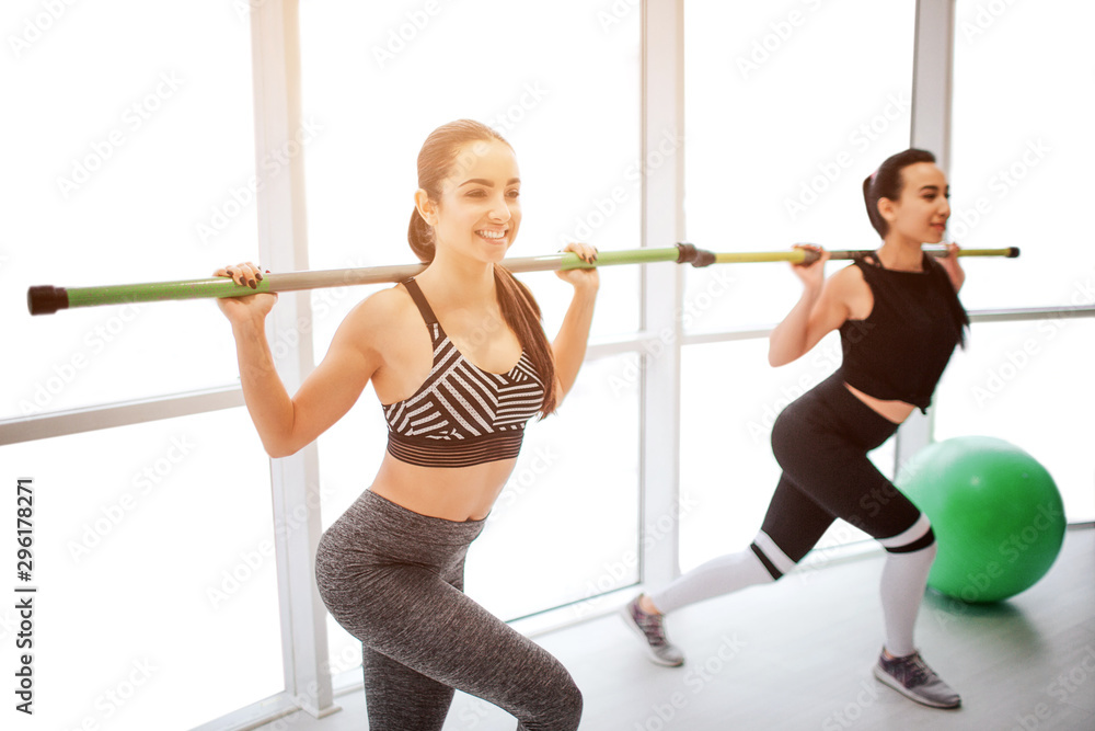 Positive and well-built young women exercising in white room. They hold barbell on shoulders and do squad moves. Models exercise together.