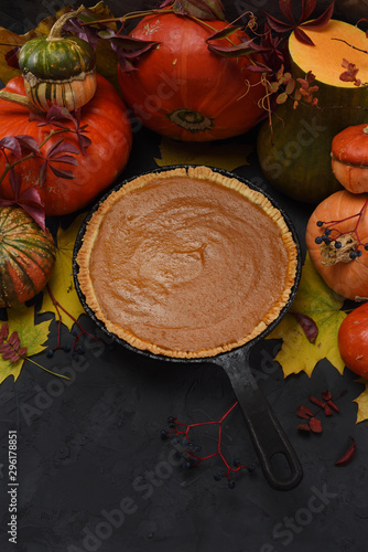 Traditional pumpkin pie with cane sugar in cast iron pan decorated with raw squashes and autumn leaves on black background copy space. Low key still life photo