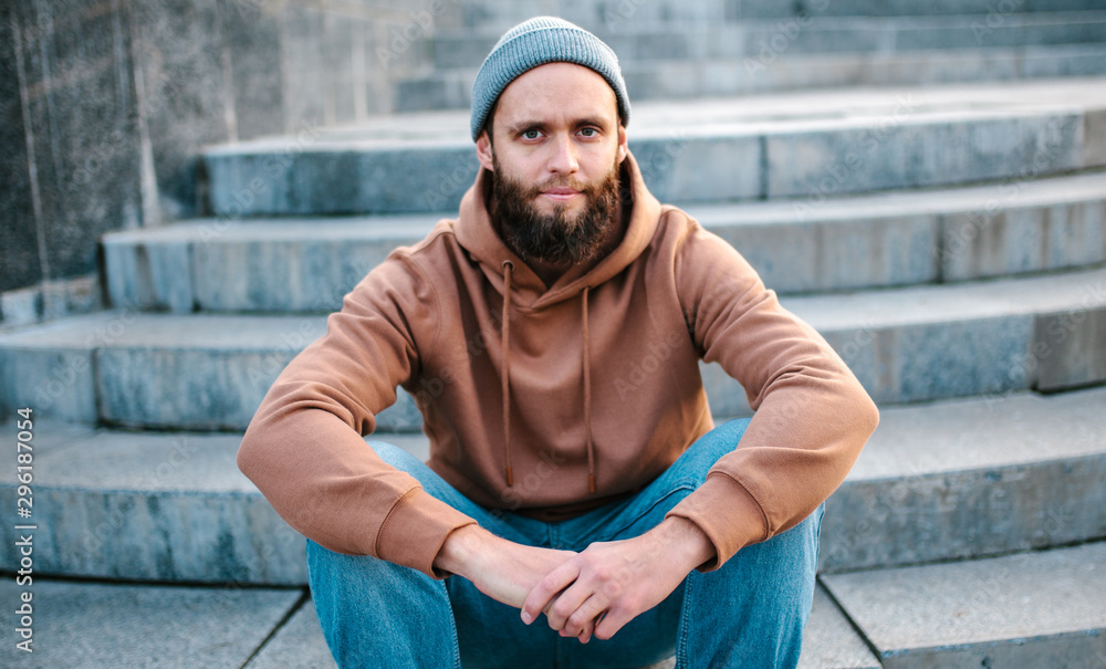 City portrait of handsome hipster guy with beard wearing brown blank hoodie or hoody and hat with space for your logo or design. Mockup for print