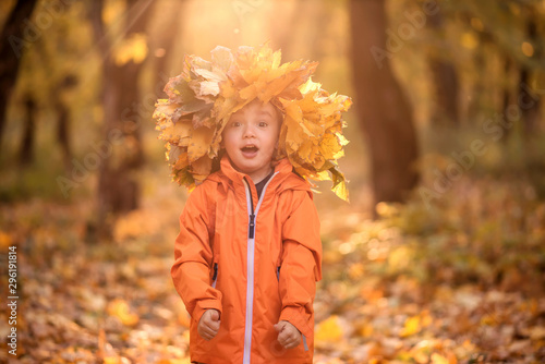 Surprised little child in crown of autumn leaves with funny face and open mouth standing in park or forest outdoors. Yellow trees in background. Seasonal fun  autumn activities and happy fall concept