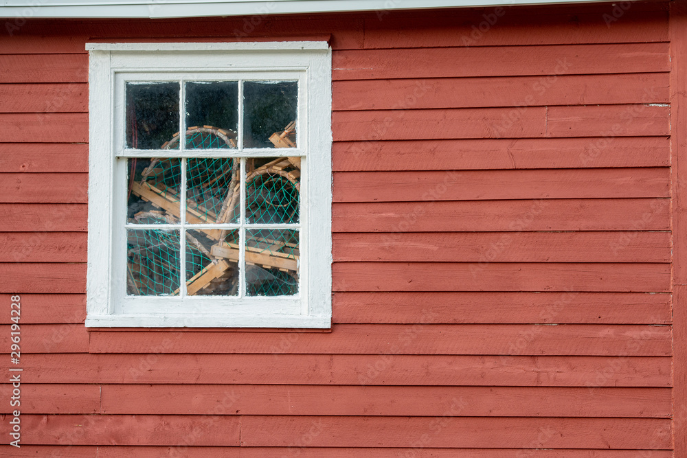 A red exterior clapboard wall with a small nine pane window with white trim. There are lobster traps in the window made of wood and green rope and netting. The old wall is textured and worn. A red exterior clapboard wall with a small nine pane window with white trim. There are lobster traps in the window made of wood and green rope and netting. The old wall is textured and worn.